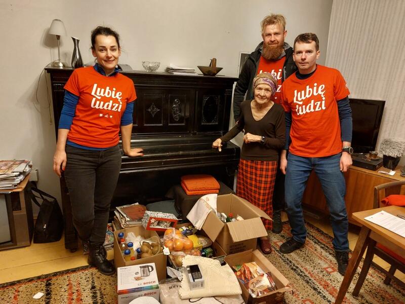 Powiększ obraz: Wolontariusze z obdarowaną Panią. Paczki z podarunkami, w tle pianino.