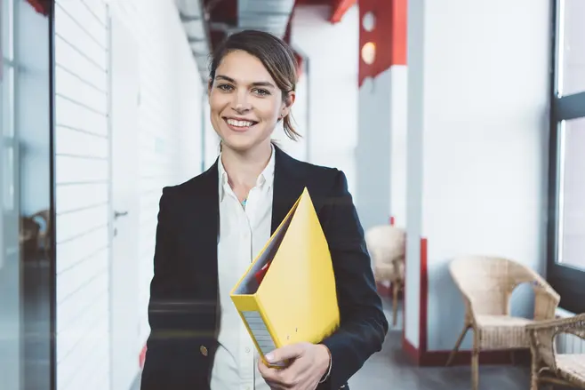 A smiling young woman in a suit, with a yellow ring binder under her arm, is walking down the corridor of an office space